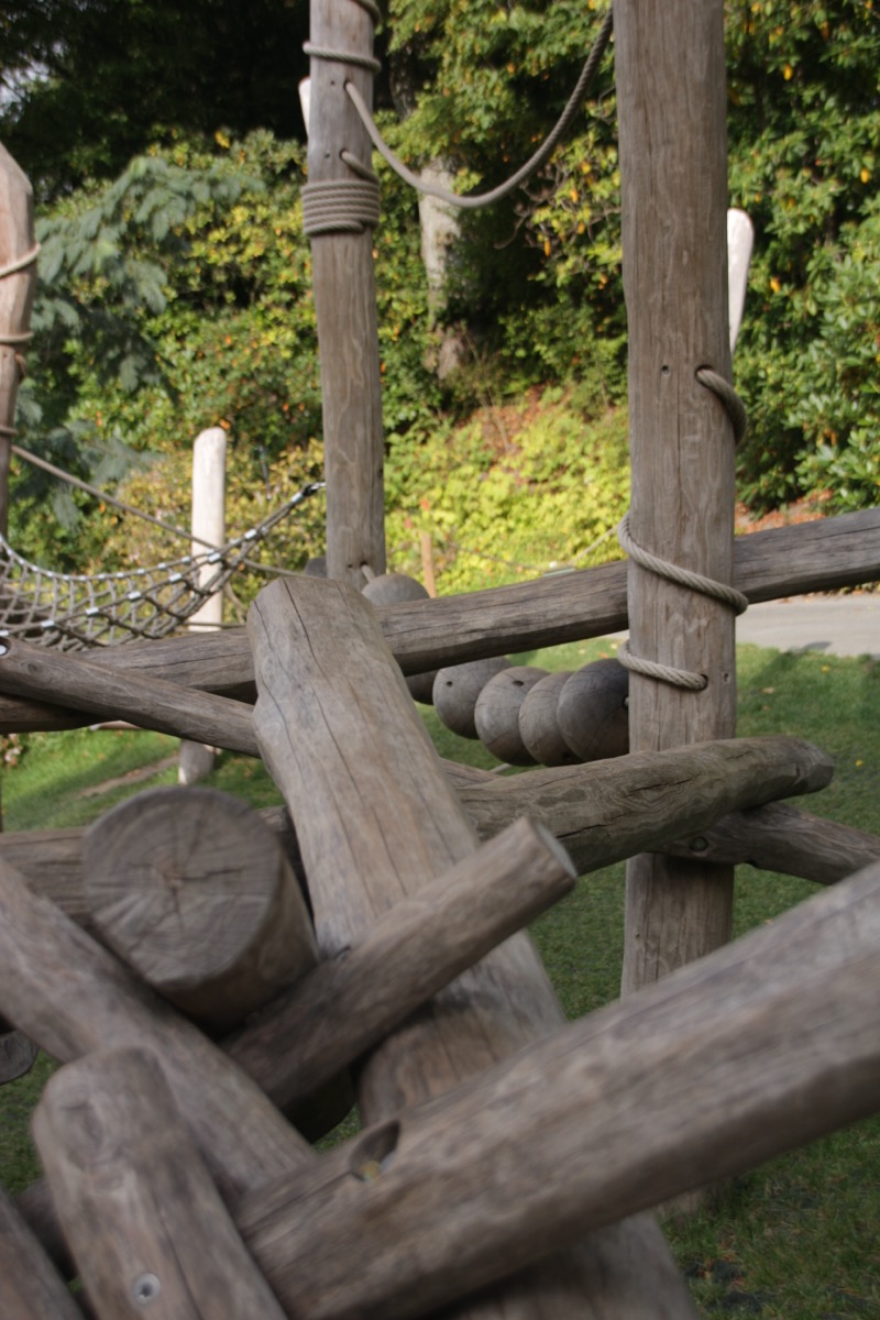 Das Foto zeigt ein Klettergerüst für Kinder zum Balancieren. Das Material ist naturbelassenes Holz. Das Gerüst hat unterschiedliche Elemente aus Baumstämmen, Kugeln und mehreren Ebenen. Seile sind als Handlauf angebracht.